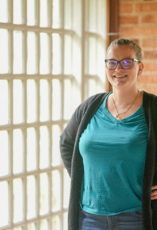 Betsie smiling at the camera near a large window. She is wearing a blue shirt and a black cardigan.