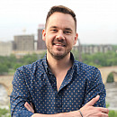 Joshua Ferchau, smiling, arms folded in a blue button-up shirt, stands in front of a city landscape.