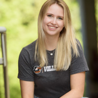 Mollee, in a Lewis & Clark volleyball t-shirt, kneeling on the ground in front of trees and smiling on a sunny day.