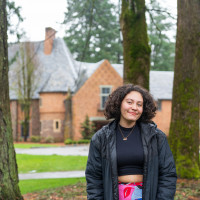 Miranda posing in front of Manor House, wearing a black jacket, black crop top, and colorful skirt.