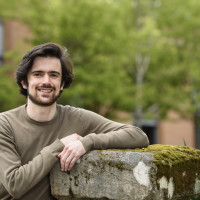 Carlos, smiling, while wearing a brown t-shirt and leaning against a concrete ledge.