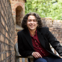 Martin, in a black blazer and red collared shirt, sitting on an outdoor bench next to a brick wall.