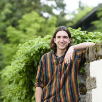 Liam, in a colorful striped collared shirt, smiling and leaning against a stone wall next to green shrubs.