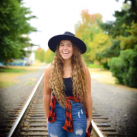 Ashtyn laughing and posing while standing on a train track, wearing a large black hat, black tank top, flannel shirt tied around waist.