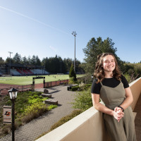 Grace posing in front of Griswold Stadium. She is wearing a black tshirt under a green jumpsuit.