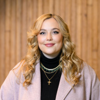 Alie smiling at the camera, wearing a pink blazer over a black turtleneck sweater.