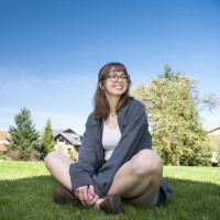 Brynn sitting crosslegged outside on a lawn. She is wearing a shite top, blue cardigan, and denim shorts.