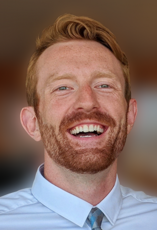Devin, in a white collared shirt and blue patterned tie, looking toward the camera and laughing.
