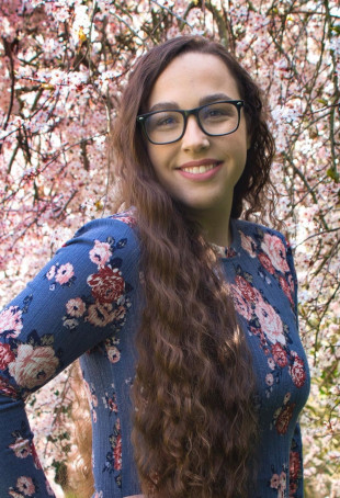 Karli smiling outside, wearing glasses and a blue long sleeve shirt with red and pink flowers on it.