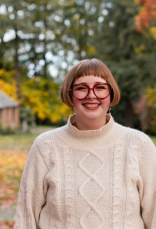 Mallory posing outside the Manor House, wearing a white sweater and red glasses.