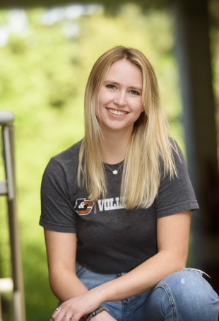 Mollee, in a Lewis & Clark volleyball t-shirt, kneeling on the ground in front of trees and smiling on a sunny day.