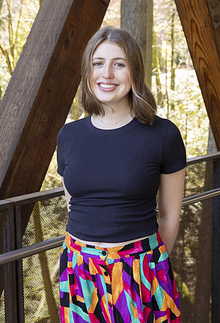 Helena smiling outside on the ravine bridge. She is wearing a black tshirt and floral skirt.