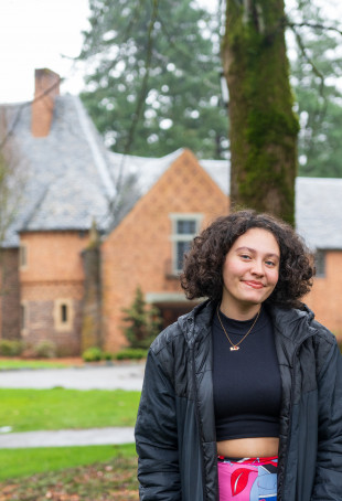Miranda posing in front of Manor House, wearing a black jacket, black crop top, and colorful skirt.