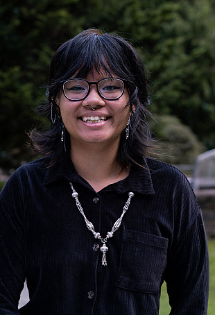 Deenie posing outside wearing a black long-sleeved shit and silver necklace.