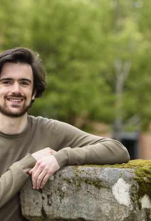 Carlos, smiling, while wearing a brown t-shirt and leaning against a concrete ledge.