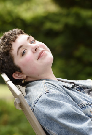 Haley, in a denim jacket, leaning back on a wooden outdoor chair while looking toward the camera.