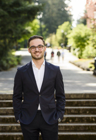 Alex, wearing a white collared shirt and navy blue blazer, smiling in front of concrete steps on a sunny day.