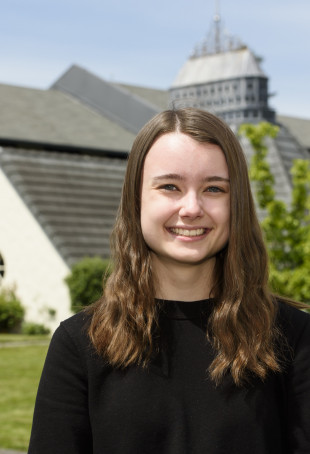 Aubrey, smiling and wearing a black long-sleeved top, standing in the courtyard of Albany Quadrangle on a sunny day.