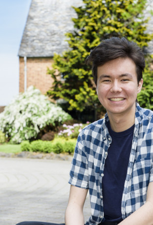 William, sitting on the ground in front of the Manor House, smiling and wearing a blue plaid collared shirt with a blue t-shirt and jeans.