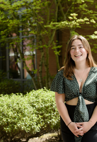 Lucia, smiling and standing in front of JR Howard Hall, wearing a green and white polka dotted top with a black skirt.