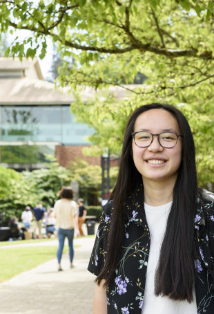 Natalie, wearing a black and floral print buttoned shirt with a white t-shirt, smiling while standing in front of Watzek Library on a sun...