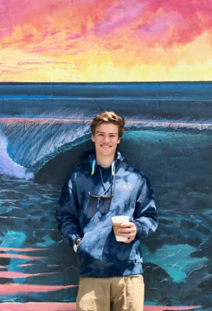 Max, holding a cup of coffee, standing in front of a wall with a painted mural of the ocean. Student-supplied profile photo due to COVID-...