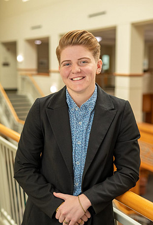 Megan posing by a stairwell, wearing a blue button-up shirt and a dark blazer.