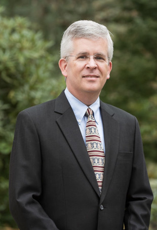 David posing outside. He is wearing glasses, a dark suit with a light shirt, and a red tie.