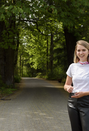 Elana, in black pants and a white t-shirt, smiling while standing in the tree-lined pathway between Flanagan Chapel and Pamplin.