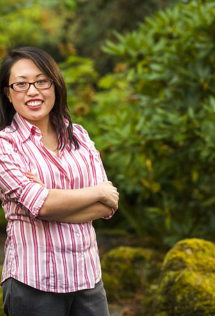 Sharon smiling outside, wearing glasses, a pink and white striped button-down shirt, and gray pants.