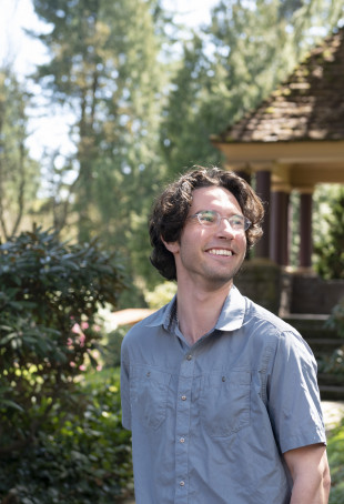 Cooper posing outside a gazebo on the undergrad campus. He is wearing glasses and a blue button-down shirt.