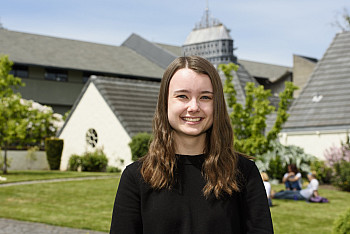 Aubrey, smiling and wearing a black long-sleeved top, standing in the courtyard of Albany Quadrangle on a sunny day.
