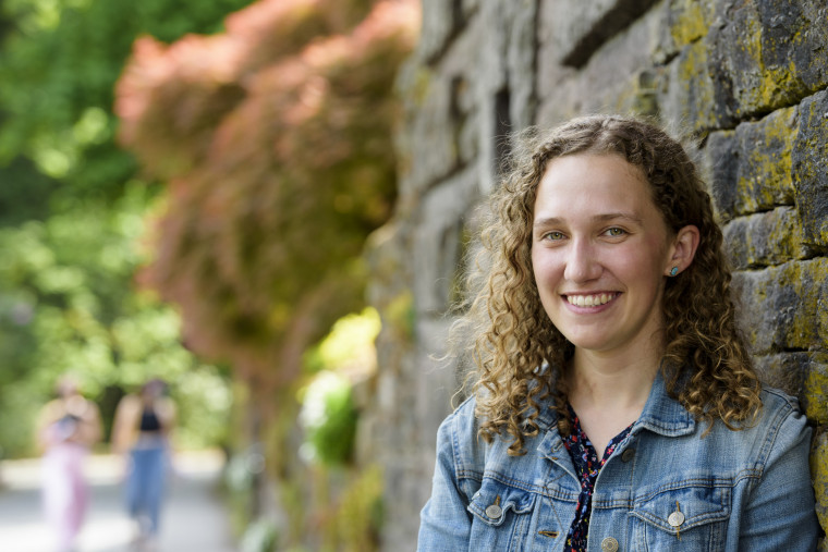Mara, wearing a floral print shirt and denim jacket, smiling in front of a stone wall near the campus reflecting pool.