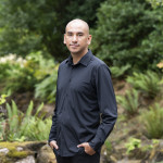 Emmanuel Macias smiling, wearing black shirt, forest background.