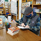 River, a transmasculine person wearing a flatcap, glasses, and a mask over their mouth and nose is seated at a table looking down at the ...