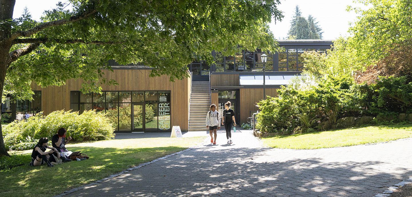 Students walking outside on a pathway with a large building behind them. There are lots of green trees and bushes and a green lawn.