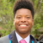 Vasty wearing a grey blazer, white shirt and pink tie, smiling with greenery in the background.