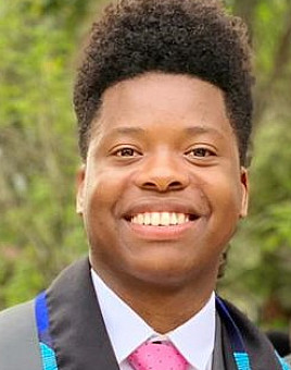 Vasty wearing a grey blazer, white shirt and pink tie, smiling with greenery in the background.