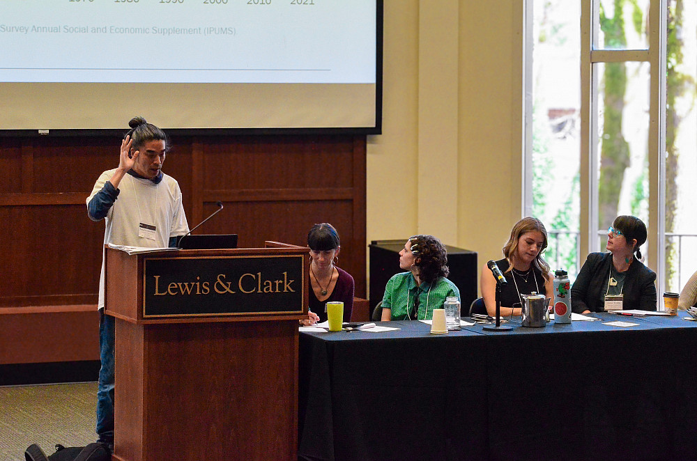 4 panelists seated at table, one speaking at podium