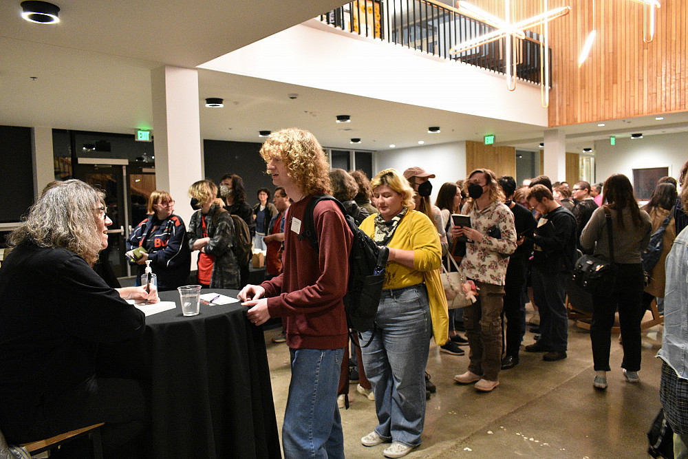 Woman seated at table, with people lined up to see her