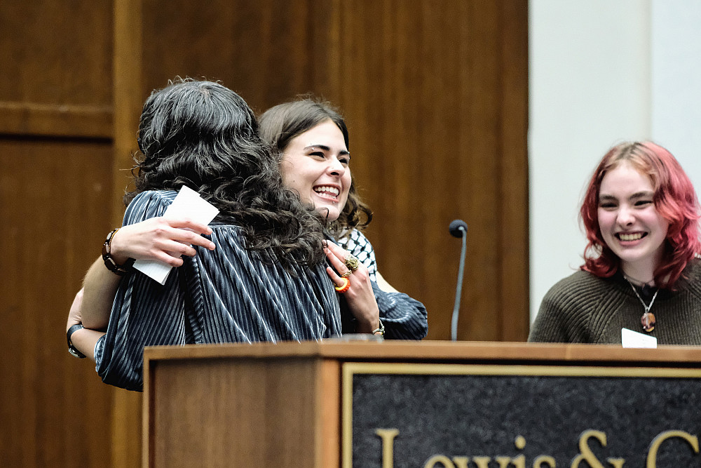 Female hugging a female student at podium while another looks on, smiling