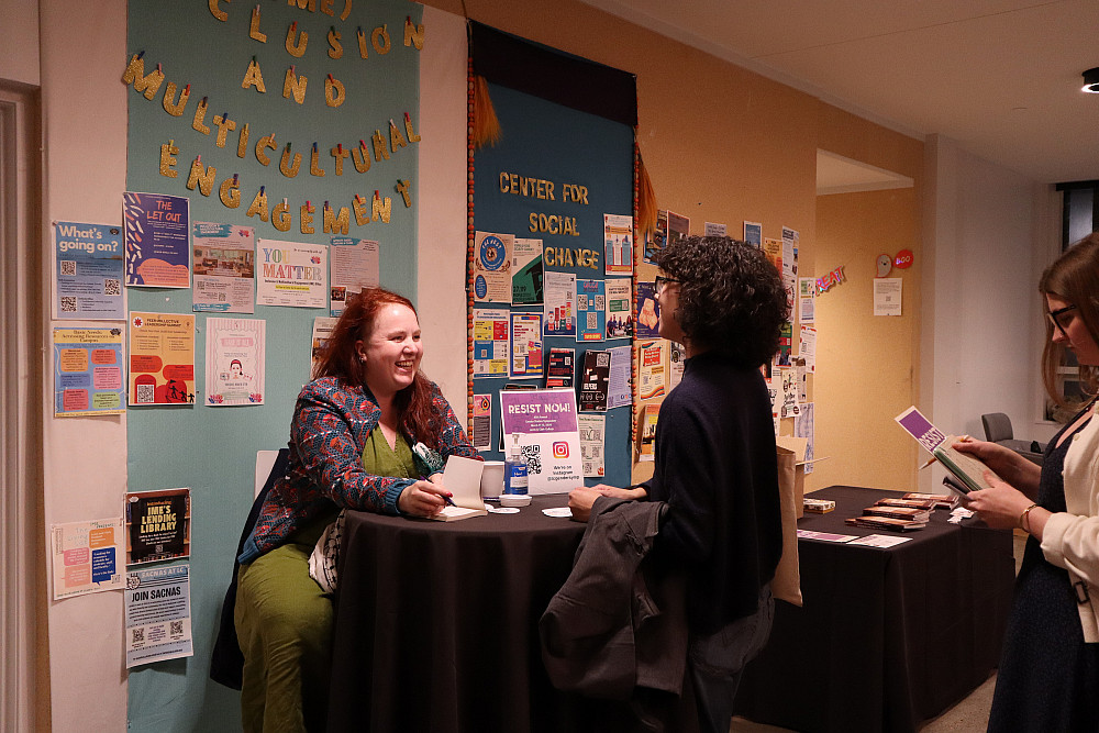 Two females seting at bistro table while one signs a book.