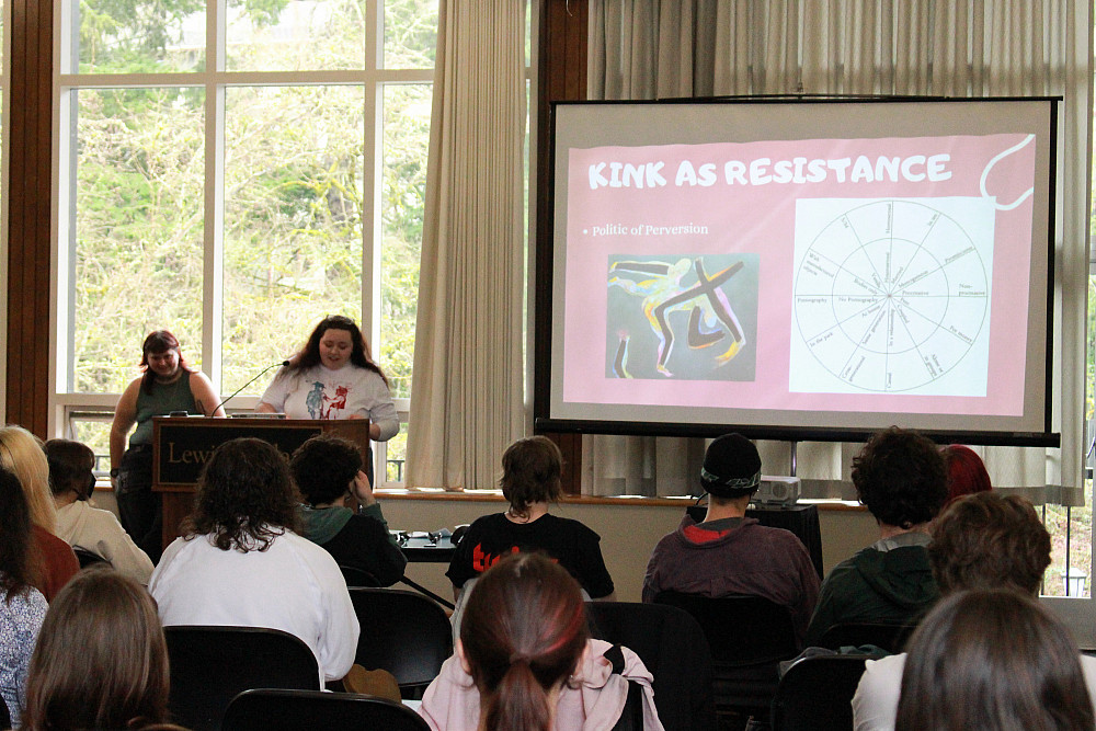 Two students at a podium, with a large screen nearby with a Kink as Reistance image, and seated students looking on.