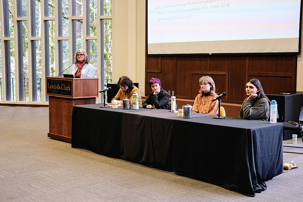 A female at a podium, next to a table with 4 seated panelists.