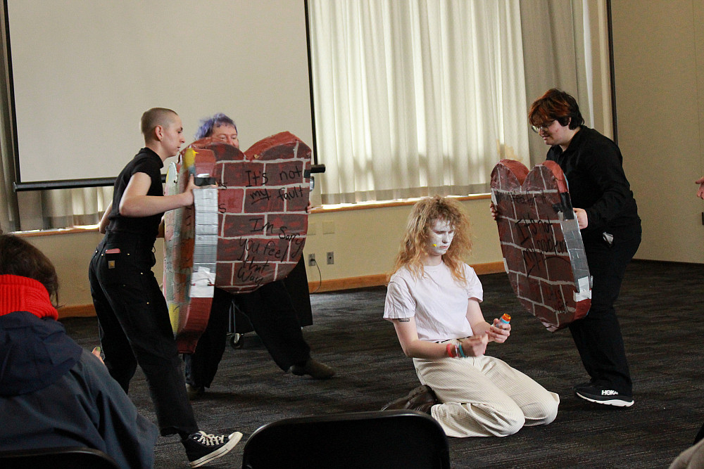 A person with white face on their knees, surrounded by 3 people holding shields painted like brick walls.