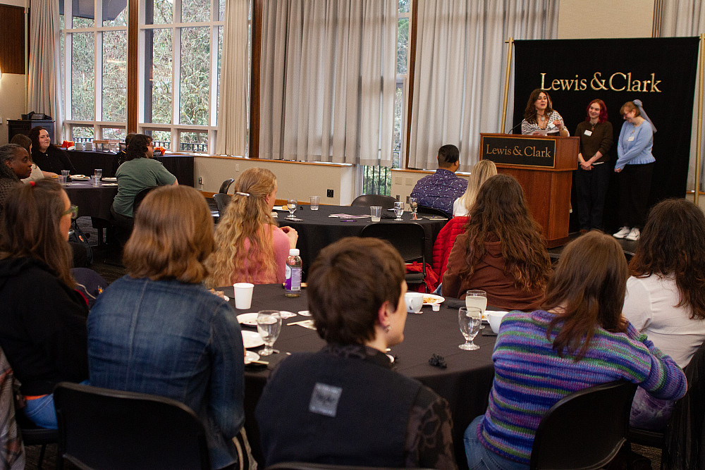 People seated in chairs at table, viewing 3 people in the front at a podium.