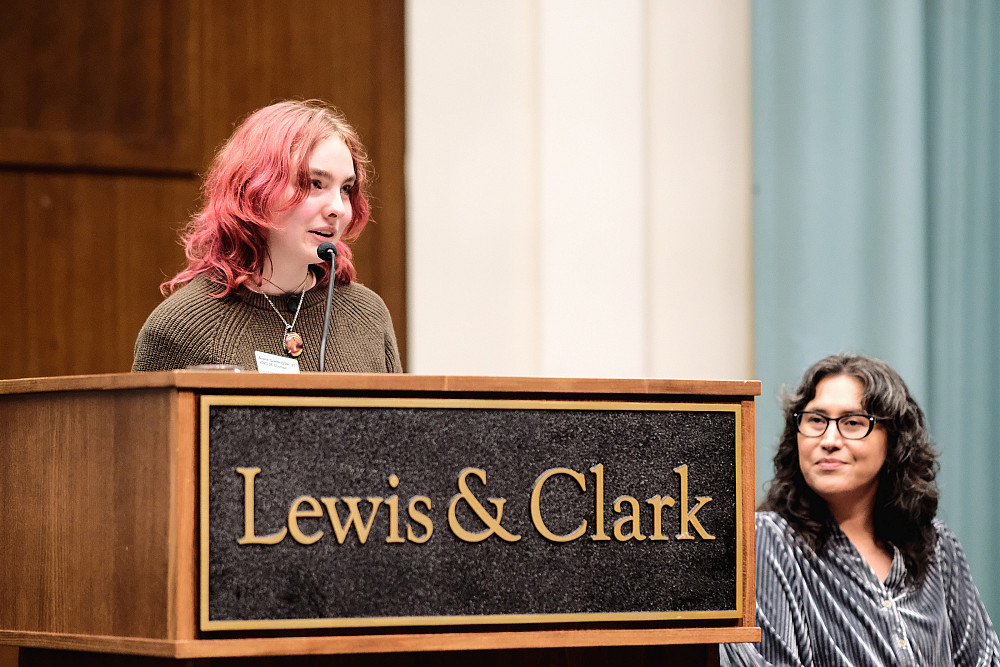 A woman speaks into a microphone at a podium while a seated woman next to her looks on.