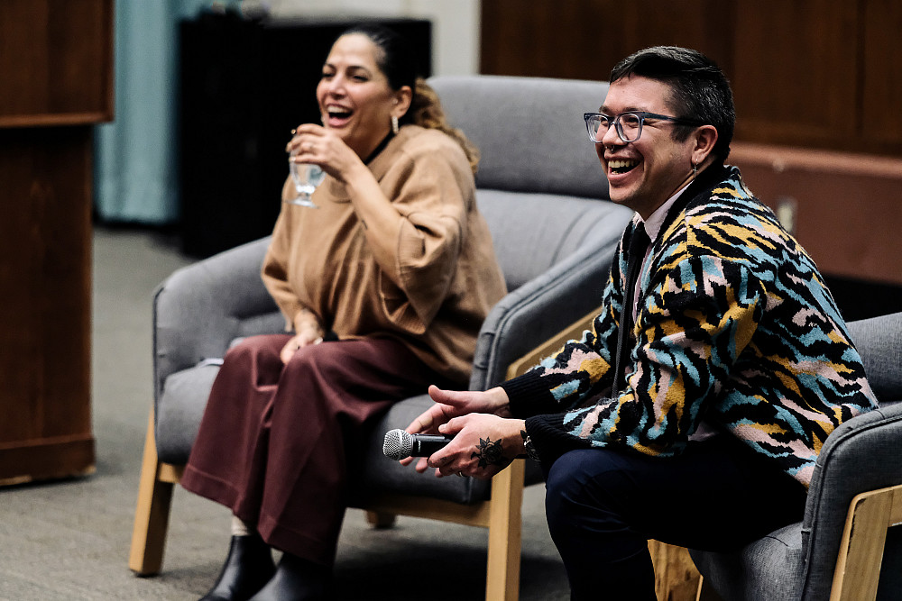 Two people in business attire seated and laughing, one holding a microphone, one holding a water glass