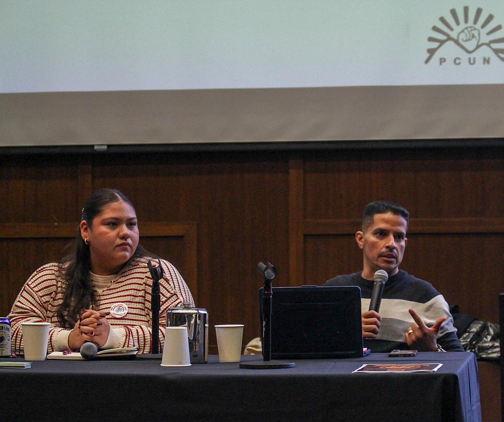Two people seated at a table with microphones, PCUN logo shown projected above them