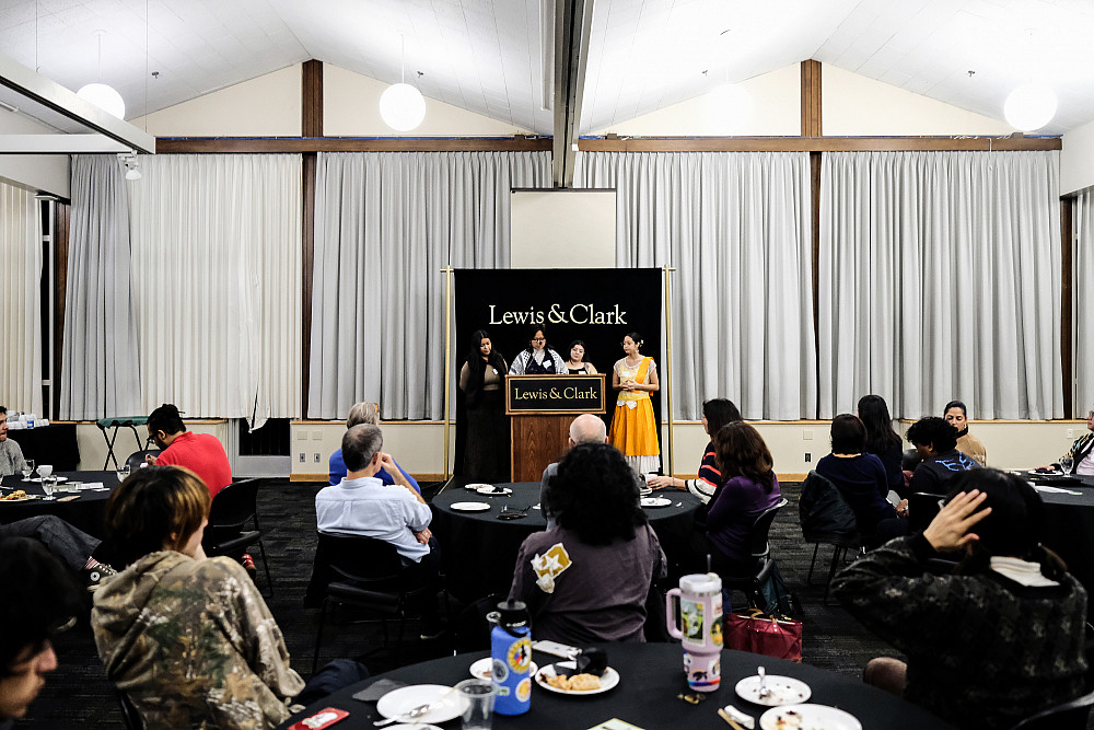 Four symposium cochair students stand at a Lewis & Clark podium in from of a backdrop on a stage, tables with audience members